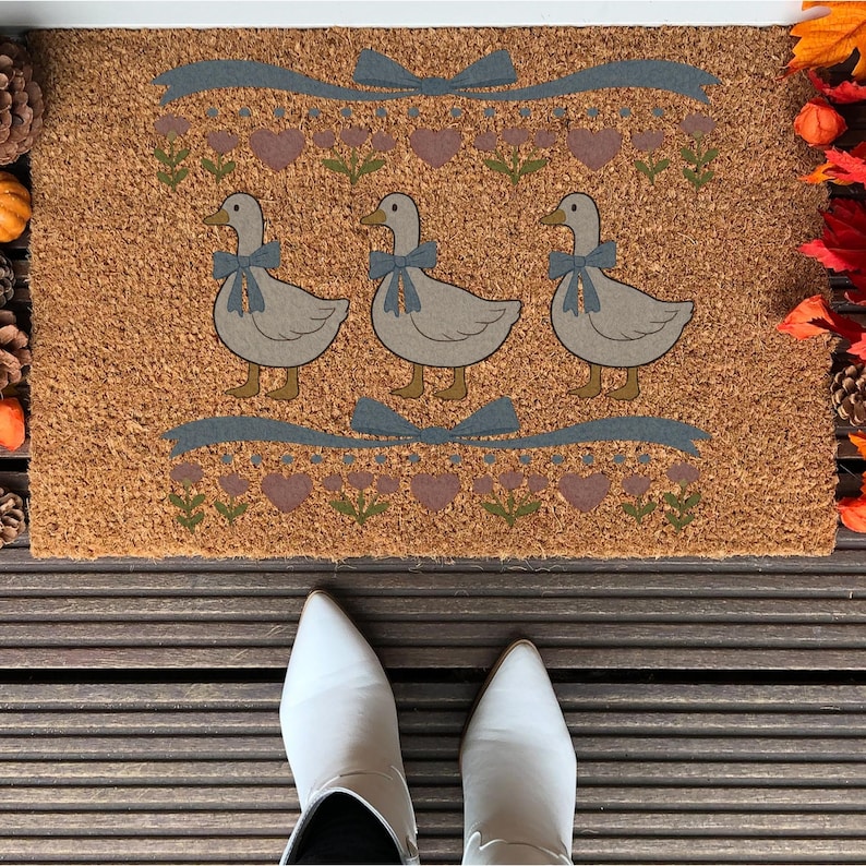 May include: A rectangular doormat with a brown coir surface. The mat features three cartoon geese wearing blue bows, with a blue ribbon and floral design above and below. The mat is decorated with hearts and flowers.