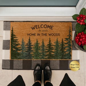 May include: A rectangular doormat with a brown center and a black and white checkered border. The center features a forest scene with green trees and the text "WELCOME to our HOME IN THE WOODS".