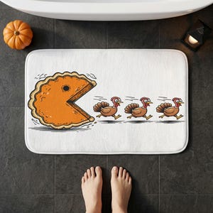 Könnte beinhalten: Eine rechteckige weiße Badematte mit einer grafischen Illustration eines großen orangefarbenen Kürbiskuchenstücks in Form einer Videospielfigur, das drei braune Truthähne in einer horizontalen Linie über die Matte jagt.