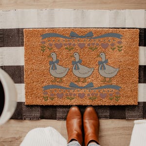 May include: A rectangular doormat with a textured, tan surface. The mat features three gray geese with blue bows, surrounded by a floral and heart design. A blue ribbon design is above and below the geese. The mat is on a black and white checkered rug.