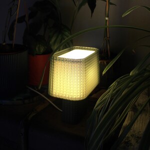 Könnte beinhalten: Eine quadratische Tischlampe mit strukturierter Oberfläche, die ein warmes, gelbes Licht ausstrahlt. Die Lampe hat ein gitterartiges Muster und einen dunklen Sockel. Sie ist von grünen Pflanzen umgeben und schafft eine gemütliche Atmosphäre. Ein dekoratives Wohnaccessoire.