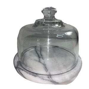 May include: A clear glass cloche with a rounded top and a small knob handle. It sits on a white marble base with gray veining. The cloche is designed to cover and protect items.