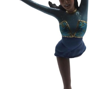 Peut inclure: Figurine d'une patineuse artistique dans une pose dynamique. La patineuse porte un costume bleu sarcelle et bleu avec des accents dorés, une jupe courte et des patins à glace. Le bras de la figurine est tendu et le visage est détaillé.