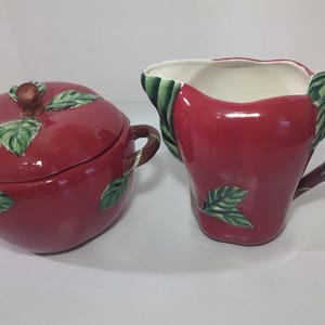 May include: A red ceramic sugar bowl with a lid and a matching creamer. Both pieces are shaped like apples, with green leaf accents. The creamer has a brown handle with green leaves.