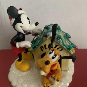 May include: A festive figurine of Mickey Mouse and Pluto. Mickey, wearing a Santa hat, decorates Pluto's doghouse with fairy lights. Pluto, with a bone and red collar, sits in front of the doghouse. The base is white and glittery.