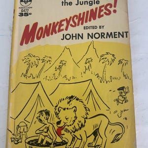 以下が含まれることがあります： ジョン・ノーメント著の「Best Laughs from the Jungle! Monkeyshines!」というタイトルのヴィンテージペーパーバック。表紙は黄色い背景に、ライオン、浴槽に入った人、ジャングルの風景の漫画イラストが描かれています。この本はバークレーメダリオン版です。