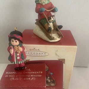 May include: Two Hallmark Keepsake ornaments. One is a girl in a red coat holding a gingerbread man and candy cane. The other is a figurine of a child sledding with a dog. Boxes read "Keepsake Ornaments" and "25 Years of Collecting Memories 1973-1998."
