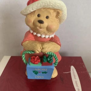 May include: A vintage Hallmark Christmas ornament featuring a teddy bear wearing a red dress, a white hat with a red accent, and a pearl necklace. The bear holds a blue box with the word "MOM" and the year "1993" on it. The box contains two wrapped gifts.