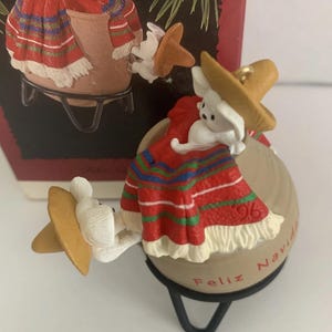 May include: A Christmas ornament featuring a white mouse wearing a sombrero and a red, white, and blue serape. The ornament is inscribed with "Feliz Navidad" and the year "96". The mouse is perched on a beige ball.