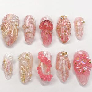 Può includere: Una collezione di dieci unghie finte con vari design. Le unghie presentano una gamma di colori rosa, bianco e oro, con abbellimenti come perle e glitter. Alcune unghie hanno elementi 3D, tra cui disegni di meduse e conchiglie.