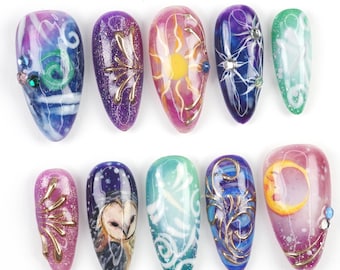 Op de afbeelding: Een verzameling van tien kunstnagels met diverse ontwerpen. De nagels tonen een scala aan kleuren, waaronder blauw, paars, roze en groen, met ingewikkelde patronen, gouden accenten en kleine versieringen. Sommige nagels beelden hemelse thema's uit zoals zonnen, manen en sterren.