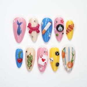 Puede incluir: Un conjunto de diez uñas artificiales en forma de almendra, cada una con un diseño único. Los diseños incluyen un pez azul, un perro globo rosa, un cigarrillo, una bola 8, un cruasán, una cereza, una serpiente, una carta de juego, dados y naranjas.