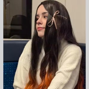 May include: A person with long, dark hair with orange tips, styled with a light pink bow. They are wearing a white, textured sweater. The background is a blurred interior with a blue seat.
