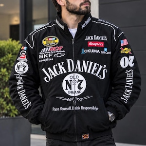 以下が含まれることがあります： 黒いジップアップのレーシングジャケット。白いジャックダニエルのOld No. 7ロゴ、NASCARネクステルカップシリーズ、スナップオン、オークマ、GMカードのロゴが特徴です。ジャケットには「Pace Yourself. Drink Responsibly」というテキストと、様々なスポンサーのワッペンが施されています。