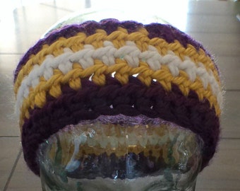 May include: Purple and gold crocheted headband with white stripes. The headband is on a glass mannequin head.