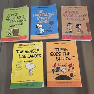 May include: Five vintage Peanuts Parade books are displayed, each with a unique cover design. The books feature titles like "A Kiss on the Nose Turns Anger Aside" and "The Beagle Has Landed," with illustrations of Snoopy and other characters.