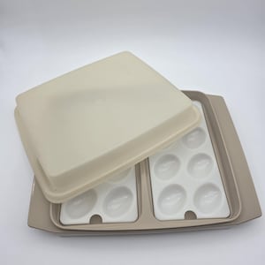 May include: A rectangular beige plastic egg storage container with a matching lid. The base features two white removable inserts, each containing six molded oval indentations designed to hold eggs securely in place within the tray.