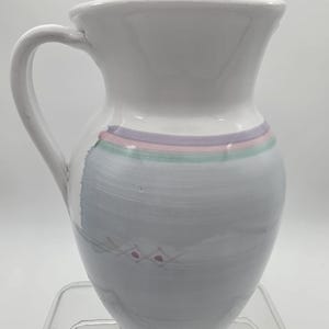 May include: White ceramic pitcher with a curved handle. The pitcher features a gray painted design with pink accents and horizontal stripes of purple, pink, and green near the top. A decorative piece for the kitchen or dining room.