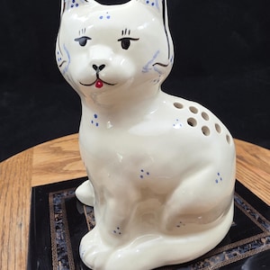 May include: A white ceramic cat figurine with black and blue accents. The cat has a red mouth, black eyelashes, and blue spots. The figurine has several holes on its back and is sitting on a decorative black surface.