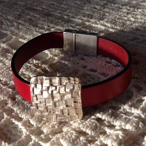 May include: A red leather bracelet with a silver square-shaped charm. The charm has a textured, geometric design.