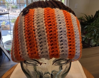 May include: A handmade crochet beanie in a pumpkin-inspired color scheme. The hat features alternating vertical stripes of orange, white, and a dark brown band at the top. The hat is displayed on a glass head.