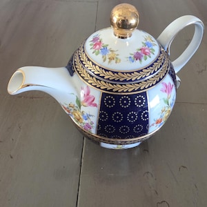 May include: White porcelain teapot with a gold finial and handle. The lid and body feature floral patterns and a navy blue band with gold accents. The teapot has a spout and is designed for serving tea.