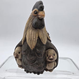 May include: A detailed, hand-carved figurine of a hen with three chicks. The hen is primarily dark brown with tan and black accents. The chicks are a light tan color. The figurine is likely made of wood or a similar material.