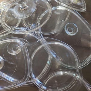 May include: A collection of clear glass serving dishes and lids. The dishes have various shapes, including round, oval, and divided sections. Each lid has a knob handle. The glass is transparent, allowing a view of the surface beneath.