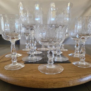 May include: A collection of clear, etched glass goblets and wine glasses arranged on a wooden serving tray. The glasses feature delicate floral patterns and are displayed on a round, wooden tray. The stemware is ideal for serving beverages.