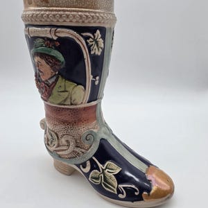 May include: A ceramic boot-shaped stein with a detailed relief design. The stein features a portrait of a man in traditional attire, along with floral and leaf motifs. The boot is primarily navy blue, with accents of tan, green, and gold.