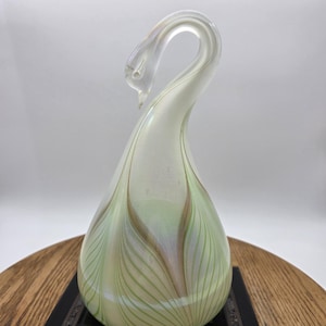 May include: A teardrop-shaped glass sculpture of a swan with a long, curved neck. The body features an iridescent finish with delicate, light green feather-like patterns and subtle gold-toned accents that swirl upward toward the swan's head.