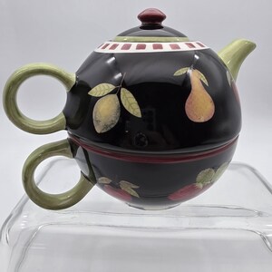May include: Black teapot and teacup set with fruit illustrations. The teapot has a red knob and a green handle and spout. The teacup is stacked below the teapot. The set is decorated with lemons, pears, and strawberries.