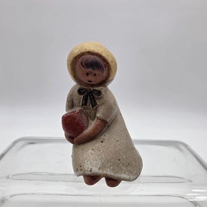 May include: A small ceramic figurine of a person wearing a speckled beige bonnet and a matching dress with a dark brown bow at the neck. The figure has dark brown hair and is holding a solid red, heart-shaped object in front of their chest.
