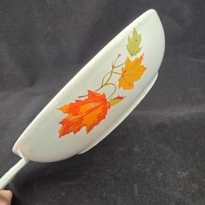 May include: A white spatula with a leaf design in orange, yellow, and green. The spatula has a long handle and a curved, flat head. The leaf design is on the head of the spatula.