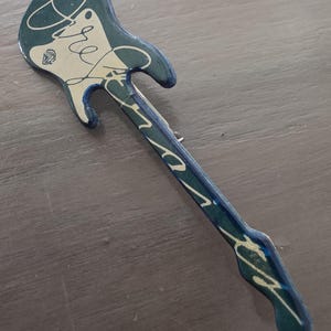May include: A guitar-shaped brooch in shades of teal and cream. The brooch features handwritten script and a diamond-shaped graphic. The design is reminiscent of a vintage or handmade item, with a slightly distressed appearance.