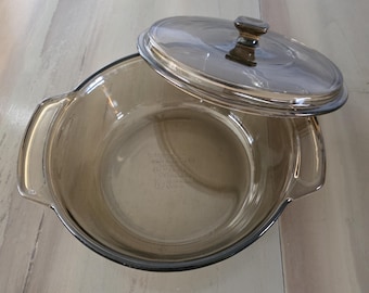 May include: A clear, round, glass baking dish with a matching lid. The dish has two handles and a dark brown tint. The lid is slightly ajar, revealing the interior of the dish. This kitchenware is suitable for baking and serving.