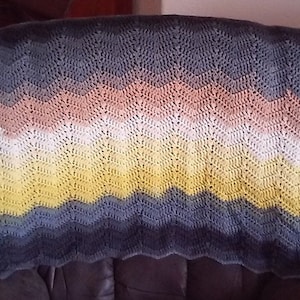 May include: A handmade crochet blanket with a chevron pattern. The blanket features a gradient of colors, including yellow, peach, white, and shades of blue and gray. The blanket is draped over a dark surface.