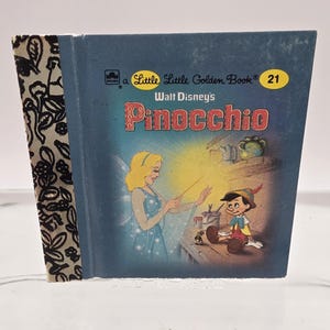 May include: A vintage Little Golden Book titled "Pinocchio" by Walt Disney. The cover is blue with illustrations of Pinocchio and a fairy. The spine has a black and white floral pattern. The book is number 21 in the series.