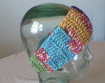 May include: A colorful, crocheted headband featuring a mix of blue, yellow, green, pink, and purple yarns. The headband is displayed on a clear glass head form, showcasing its textured stitch pattern and vibrant color palette. This accessory is perfect for adding a pop of color.