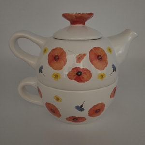 May include: White ceramic teapot and teacup set decorated with orange poppy flowers, yellow and blue wildflowers. The teapot has a red-orange lid and handle. The teacup fits neatly under the teapot.