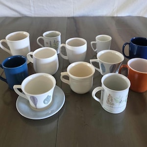 May include: A collection of ceramic mugs in various colors and designs. The mugs include solid white, blue, and orange, as well as mugs with floral and abstract patterns. Some mugs have a matching saucer.