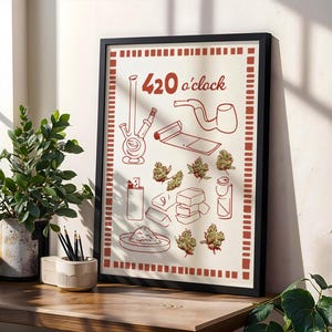 May include: A framed art print with a black border featuring a red and cream design. The print includes illustrations of smoking accessories and cannabis buds, with the text "420 o'clock" at the top. The artwork is displayed on a wooden surface.