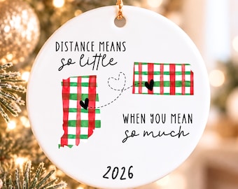 Puede incluir: Un adorno redondo de cerámica blanca con mapas de Indiana y Kansas en cuadros rojos y verdes, unidos por una línea discontinua con un corazón. El texto dice: DISTANCE MEANS so little WHEN YOU MEAN so much, con el año 2026 en la base.