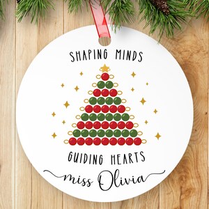 May include: A white, round Christmas ornament with a red ribbon. The ornament features a Christmas tree design made of red and green circles, with the text "SHAPING MINDS GUIDING HEARTS miss Olivia". Gold stars are scattered around the tree.