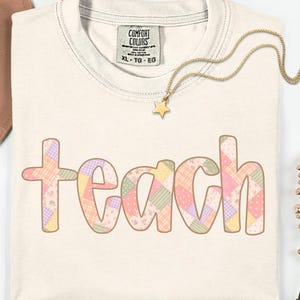Op de afbeelding: Crèmekleurig T-shirt met het woord "teach" in een patchwork-ontwerp. De letters zijn gevuld met pastelkleurige vierkanten en bloempatronen. Een gouden ketting met een sterhanger ligt op het shirt.