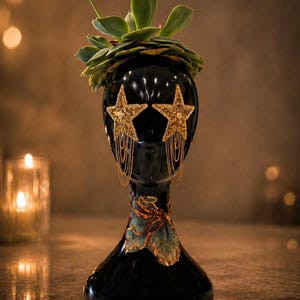 Könnte beinhalten: Ein schwarzer dekorativer Kopf mit goldenen Sternohrringen und einer Sukkulentenpflanze. Der Kopf hat ein detailliertes Blumendesign am Halsansatz. Die Ohrringe haben goldene Kettenfransen.