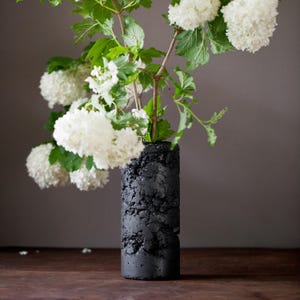 Könnte beinhalten: Eine zylindrische Vase mit einer dunkel schwarzen, stark texturierten und porösen Oberfläche. Die Vase enthält mehrere Stiele weißer Schneeballblüten mit hellgrünen Blättern, was einen Kontrast zwischen dem dunklen Gefäß und den hellen Blüten erzeugt.