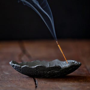 Pode incluir: Um porta-incenso circular cinzento-escuro com textura e rebordo irregular e rugoso. Uma vareta de incenso fina está colocada dentro da taça, emitindo um rasto visível de fumo branco que se eleva verticalmente contra um fundo escuro.