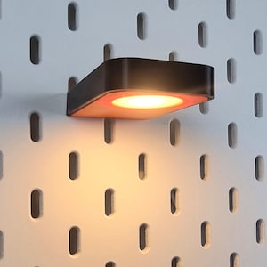 Könnte beinhalten: Eine kleine, quadratische, schwarz-orangefarbene LED-Leuchte ist an einer weißen Lochwand befestigt. Das Licht strahlt ein warmes, orangefarbenes Leuchten aus. Die Lochwand hat gleichmäßig verteilte, ovale Löcher.