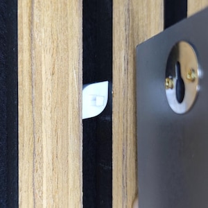 Könnte beinhalten: Nahaufnahme eines Wandobjekts mit einer schlüssellochförmigen Metallplatte und einem weißen Kunststoffclip. Die Wand weist vertikale Holzpaneele in abwechselnden hellbraunen und schwarzen Streifen auf.